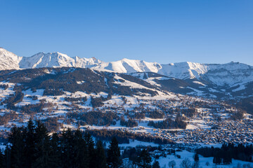 Expansive winter view of Megeve village nestled among snow-covered hills, dense pine forests, and dramatic sunlit mountain ridges under a vivid blue sky.