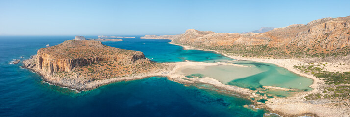 Expansive panoramic image of Balos Beach in Crete, Greece, featuring a striking turquoise lagoon, rugged rocky peninsula, and sunlit arid hills under a clear blue sky. The landscape is open and © Florent