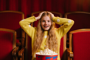 Excited young girl watching a movie in a cinema, holding popcorn and reacting emotionally while sitting in red theater seats.