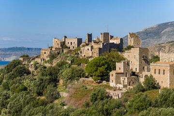 Fototapeta premium A cluster of weathered stone towers and houses rises above dense green vegetation on a sunlit hillside in Vathia, Mani, Greece. The scene features rugged textures, Mediterranean flora, and distant