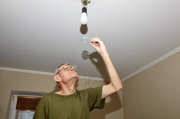An older man changing light bulb at home. He is holding a burned-out, clear incandescent light bulb in his raised right hand, inspecting it or preparing to dispose of it