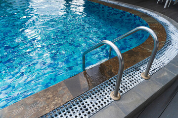 Sparkling blue water ripples in a pool featuring a stainless steel ladder. A brown stone border and white overflow grate line the edge, creating a clean and inviting summer scene.