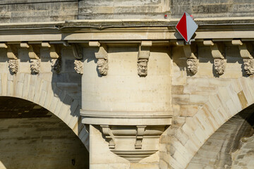Close-up of ornate stone carvings and a red navigation sign on the historic Le Pont Neuf bridge in Paris. Warm sunlight highlights the textured masonry and sculpted faces along the arch.