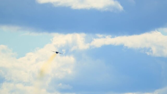 Military fighter jet flies through a cloudy blue sky, leaving a trail of colored smoke behind as it moves across open airspace in a dramatic aerial scene.