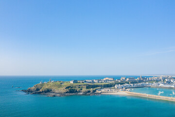 Aerial view of Granville's rugged peninsula with grassy cliffs, lighthouse, and marina surrounded by vivid turquoise sea under a clear blue sky. The coastal scene is expansive, sunlit, and serene with