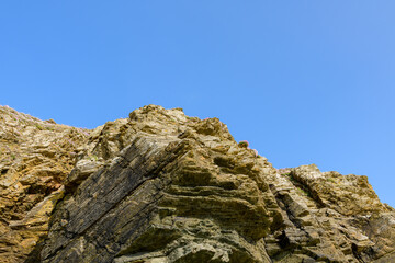 Angular rock formations rise sharply at Cap de Carteret, their textured surfaces and layered strata highlighted by bright daylight against an expansive blue sky. The rugged geology and warm earth