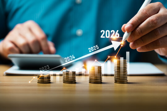 Business hand drawing upward financial growth chart with stacked coins on desk, calculator background, showing annual profit increase from 2023 to 2026 investment planning concept