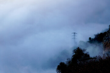 Electricity transmission pylon on a hillside rising through thick low clouds, creating a mysterious and foreboding atmosphere