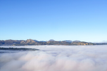 Mountain range and distant hills emerge from a thick blanket of fog or clouds, bathed in bright morning light under a clear sky
