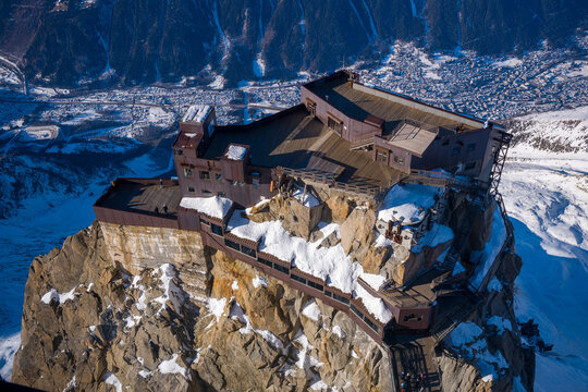 Aerial view of the Aiguille du Midi summit station perched dramatically on rugged, snow-dusted cliffs above the Chamonix valley in bright winter sunlight.