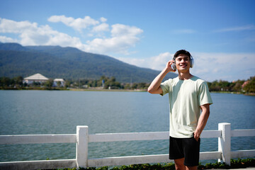Photo of young asian man with headphones rest next to a river relaxing enjoying the sun and music