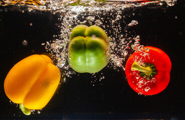 High speed underwater shot of yellow, green, and red bell peppers creating dynamic splashes against a dark background. This vibrant image represents freshness, healthy eating, 