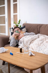 Boy wrapped in blanket using inhaler mask, sitting on sofa at home, recovering from illness, glass of water nearby, cozy indoor setting.