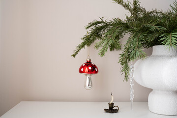 A festive glass Christmas ornament in the shape of a red-and-white mushroom hangs from a pine branch. The shiny cap with white spots contrasts with the silver stem, creating a whimsical holiday charm.
