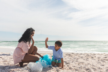Obraz premium Happy mother and son giving high-five while collecting garbage in bag at beach against sky