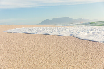 Obraz premium Sea wave foam on shore at sunny beach with mountains in distant background
