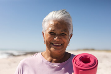 Portrait of smiling retired senior woman holding exercise mat with short hair at beach