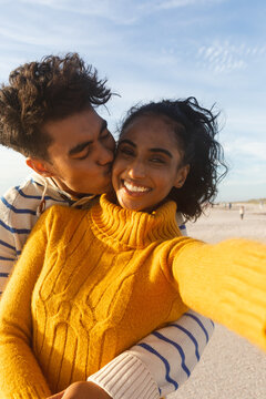 Portrait of smiling woman taking selfie of boyfriend hugging and kissing on cheek at beach