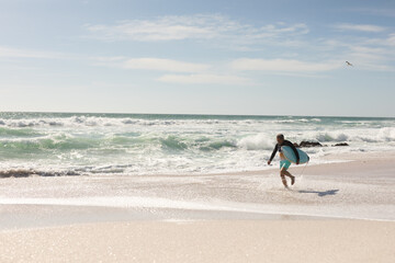 Retired senior man running with surfboard on shore at beach against sky during sunny day