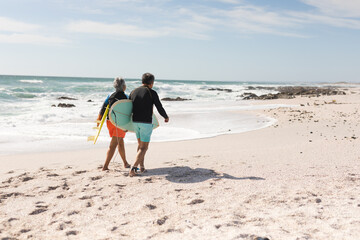 Obraz premium Rear view of multiracial senior couple walking with surfboards on shore at beach during sunny day
