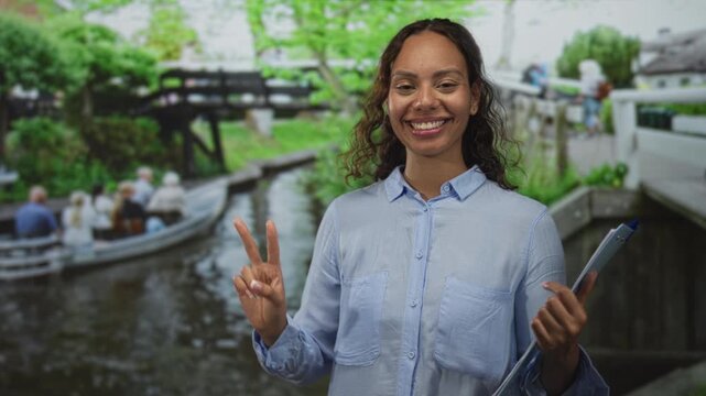 Woman smiling and showing a peace sign with her hand while holding a clipboard on an amsterdam street canal bridge; joy travel.