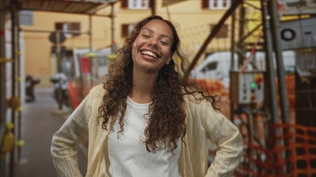 Woman laughing with eyes closed and hands on hips at building scaffolding with orange safety netting and delivery van behind; joy confidence.