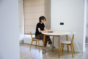 Young woman reading a book while sitting at a table in a bright modern kitchen. Calm home atmosphere, everyday routine, mindfulness and relaxed lifestyle concept.