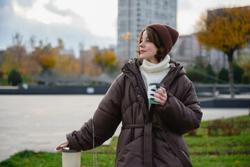 Stylish woman in a warm brown jacket and knit hat holding takeaway coffee while walking outdoors in autumn city park. Cozy seasonal lifestyle, calm mood, urban environment and slow living concept.