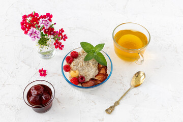 Oatmeal porridge with berries in a glass bowl and a cup of tea on the white table.