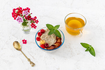 Oatmeal porridge with berries in a glass bowl and a cup of tea on the white table.