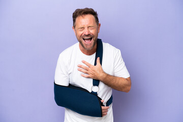 Middle age caucasian man with broken arm and wearing a sling isolated on purple background smiling...