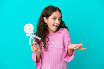 Little caucasian girl holding a lollipop isolated on blue background with shocked facial expression