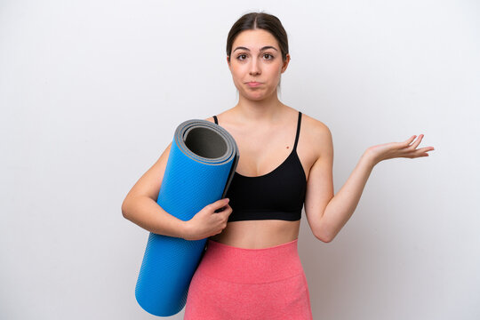 Young sport girl going to yoga classes while holding a mat isolated on white background having doubts while raising hands