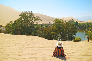 Female traveler relaxing on the sand dune, admiring the oasis town of Huacachina, Ica, Peru, South...