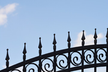 The upper part of a black wrought iron fence featuring ornate swirls and pointed spikes stands against a light blue sky with soft white clouds