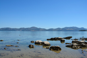 A serene landscape showing the rocky shore of Drace on Peljesac peninsula looking across the Malo More towards the mountains of mainland Croatia under a clear sky