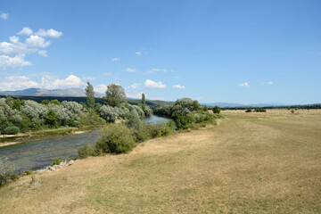 A wide landscape view captures the Cetina River winding through green trees and vast golden fields under a clear blue sky outside the village of Panj.