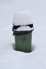 A green public trash bin stands surrounded by deep white snow with a thick snowy cap on its lid in a winter park