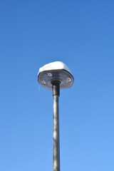 A modern street lamp is covered with a thick cap of white snow and a small icicle against a clear blue winter sky