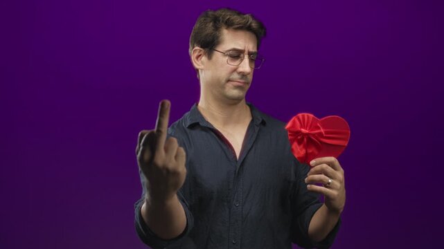 Man in dark shirt holds a red satin heart gift with bow while showing middle finger toward camera in purple studio; defiance rejection.