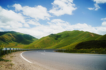 Obraz premium Winding asphalt road through lush green mountains under a bright blue sky with fluffy white clouds
