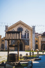 A wooden wishing well with a block sits in a paved courtyard against a backdrop of a decorative stone building under a blue sky in Armenia