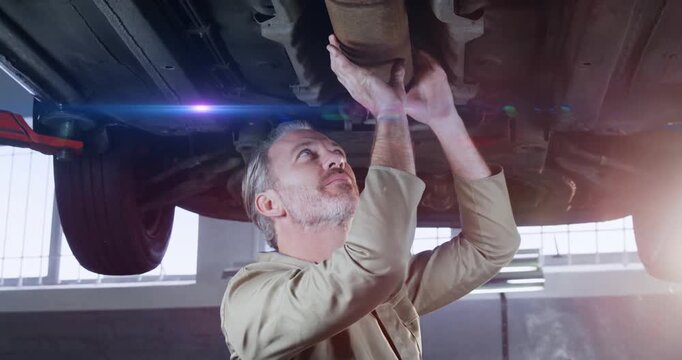 Senior male mechanic inspecting exhaust beneath car in automotive bay, showing floating data icons