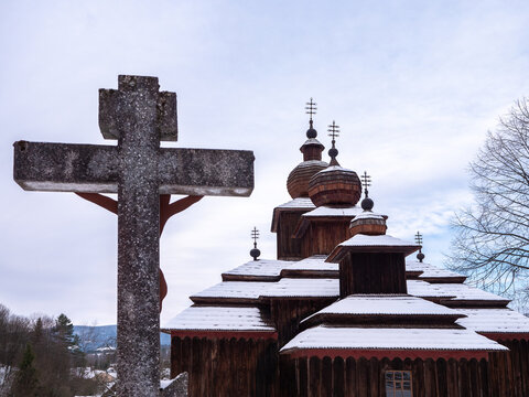 Stone cross in front of the Wooden Greek Catholic Church of St Paraskeva in Dobroslava, Slovakia. Traditional Carpathian wooden church photographed in winter with snow.