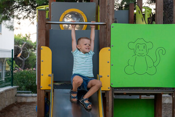 Happy smiling boy hanging on bar above metal slide on colorful playground with monkey drawing in...