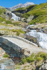 Verfical view of an ancient bridge made of stone on the path leading to "San Grato" lake, Valgrisenche, Aosta Valley, Italy, with a mountain creek full of water. High mountains peaks on the background