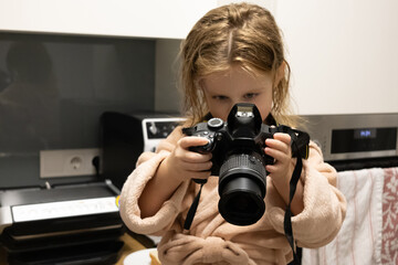 Naklejka premium Curious little girl in a beige bathrobe holding a professional DSLR camera and taking pictures in the kitchen