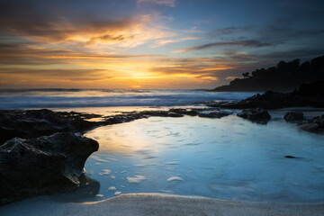 Obraz premium A golden sun setting over a rocky shoreline on an Indonesian island. Long exposure capture on sunset featuring a shallow blue tidal pool below vibrant orange clouds.