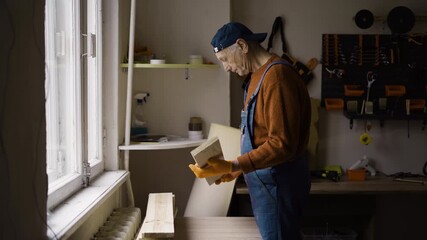 Elderly man carpenter with wooden products at workshop manufacturing furniture working in wood warehouse industry.