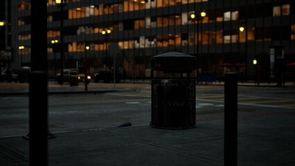 The scene captures a quiet urban street at dusk, where city lights reflect on tall buildings. A lone trash bin stands on the sidewalk, hinting at the end of the day.